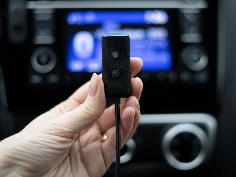 Yaklaşık 1 inç genişliğinde ve 2 inç uzunluğunda küçük siyah bir cihaz olan Echo Auto'yu tutan bir elin yakın çekimi. Küçük bir düğmesi ve bir gösterge LED'i ve ondan aşağı inen bir kablosu vardır. Fotoğraf bir arabada çekilmiştir ve orta gösterge paneli / bilgi-eğlence alanı arka planda odak dışındadır.