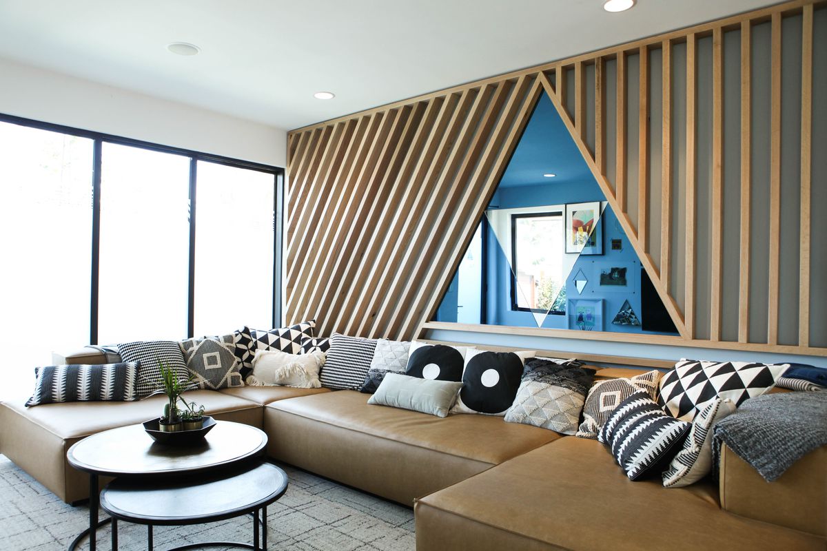 Living room with a triangular mirror, wood slats on the walls, patterned pillows and a beige couch.