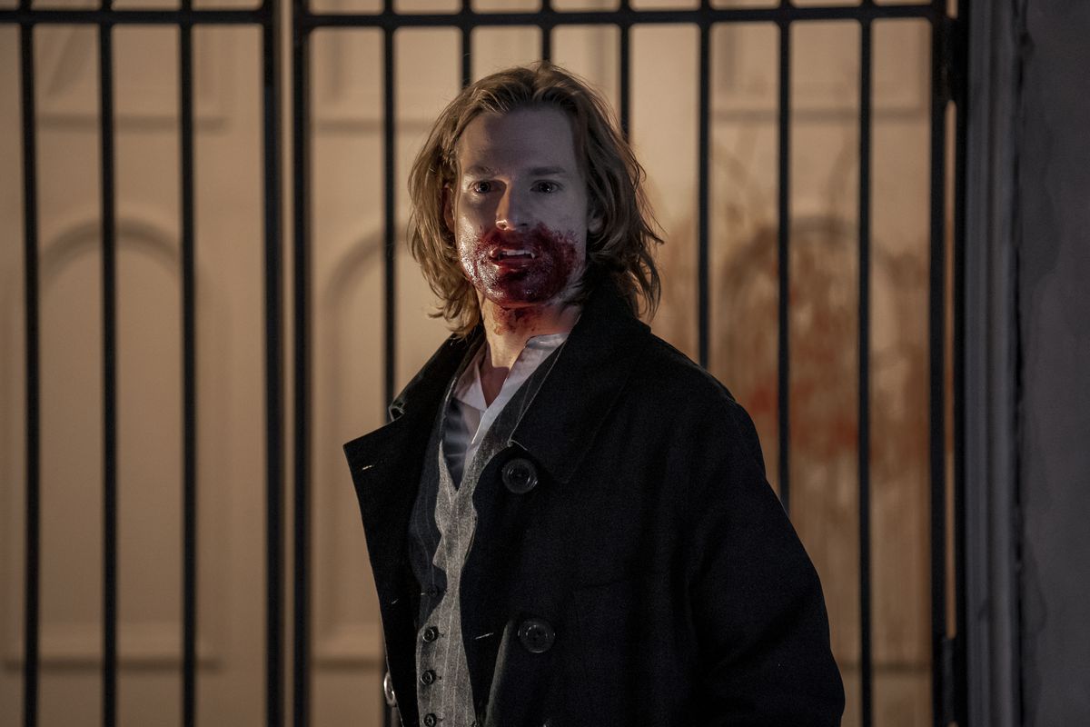 The vampire Lestat stands in an open coat with a bloody mouth in front of a wrought iron fence in AMC’s Interview with the Vampire