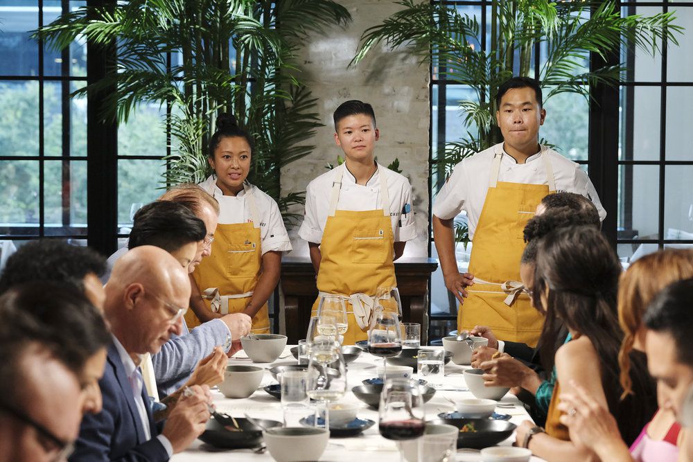 Three people in white chef coats and yellow aprons standing behind a table of people eating.