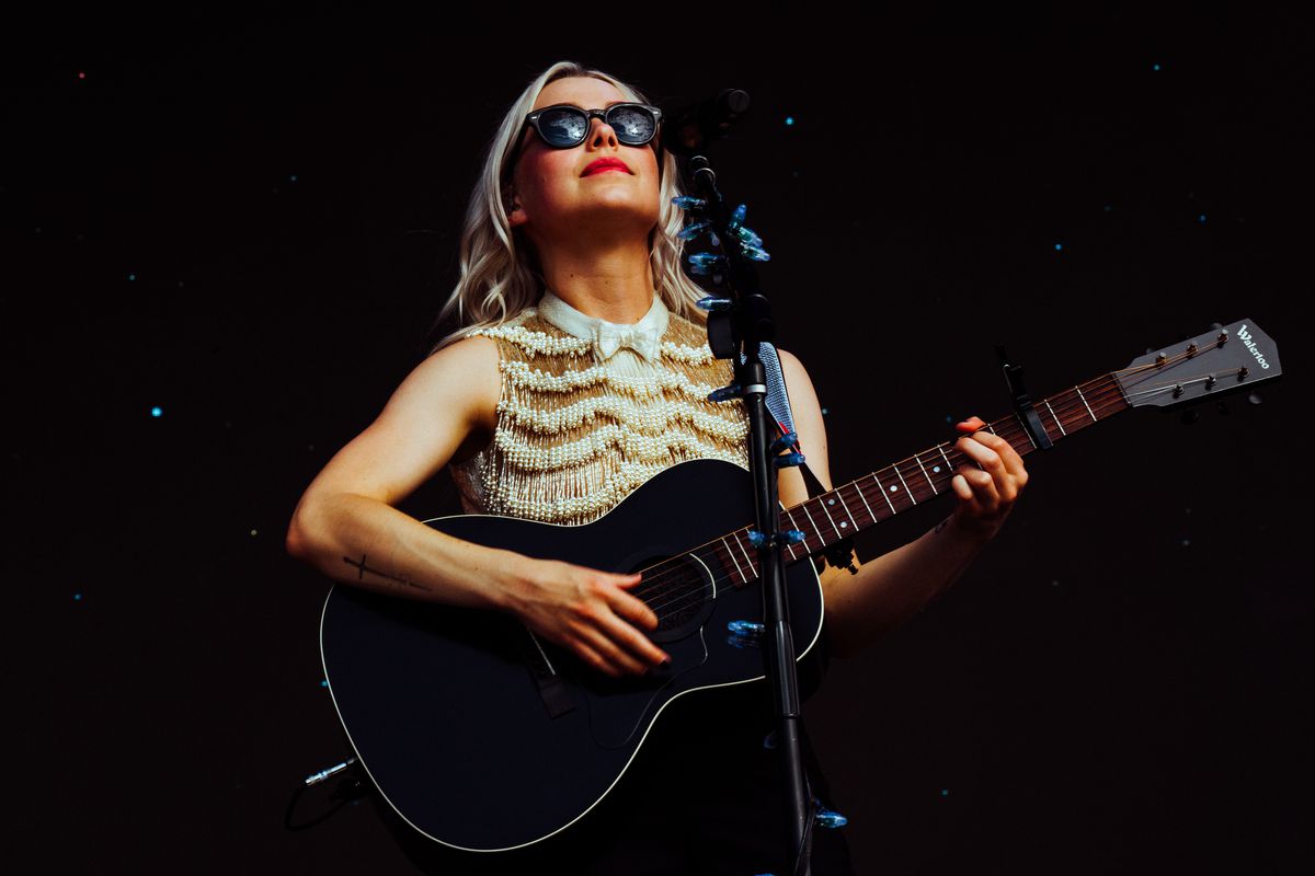A woman in a fringe shirt and sunglasses playing a guitar.