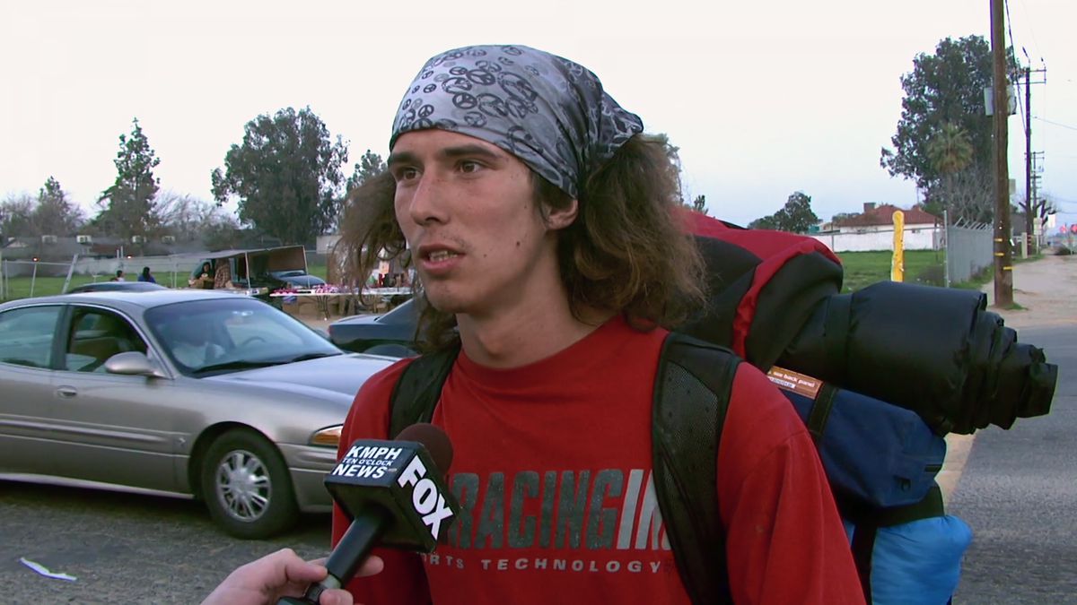 A long-haired man in a red shirt wearing a gray bandanna on his head and a backpack speaking to a microphone in a parking lot.