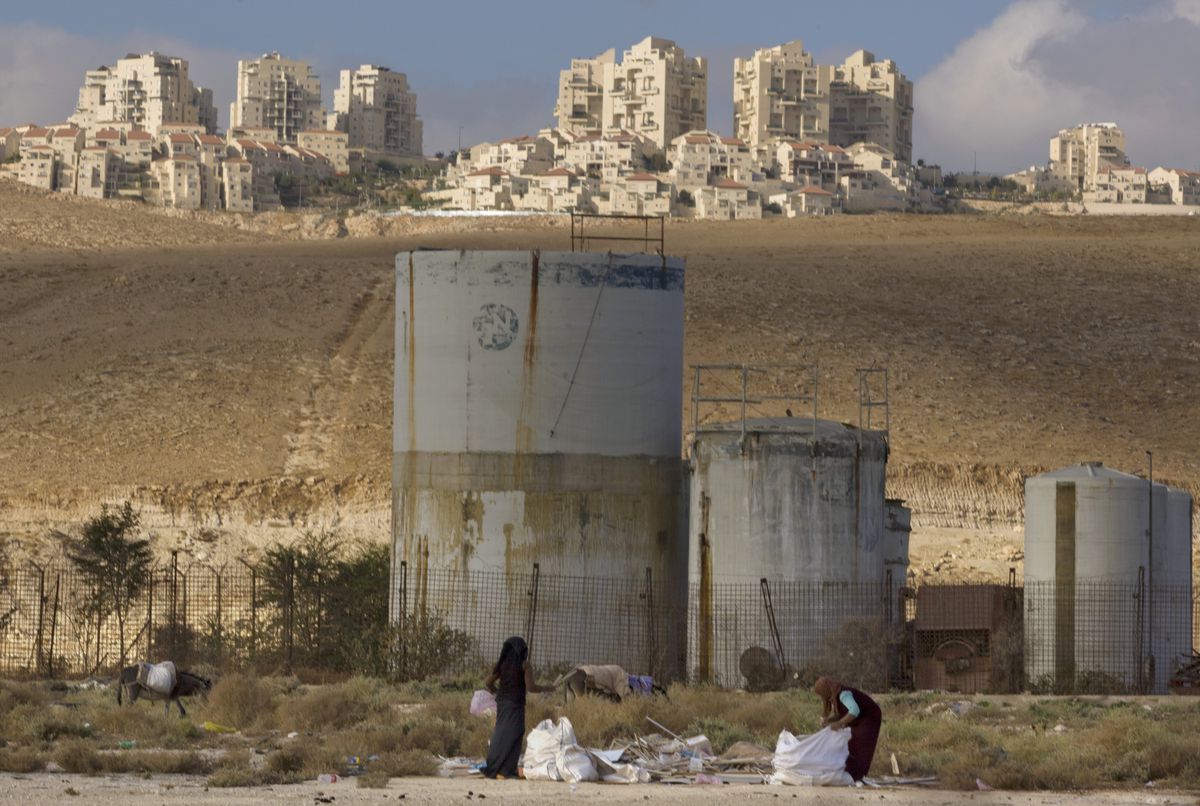 Women surrounded by dry, barren brown land, fill white trash bags. Behind them stand some industrial silos, and farther in the distance, a settlement of multistory buildings can be seen. 
