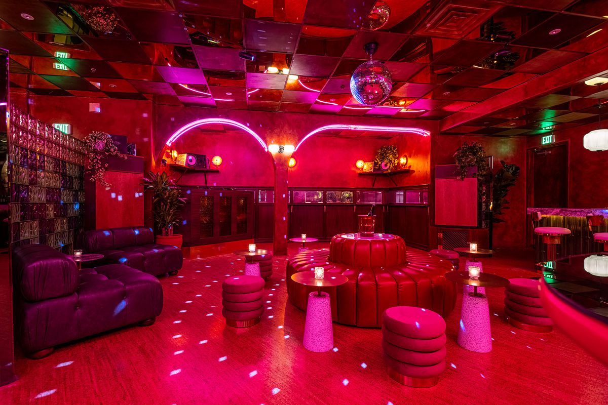 A red-hued room with a disco ball on the ceiling shining sparkles of light throughout the room that has neon lights and round couches.