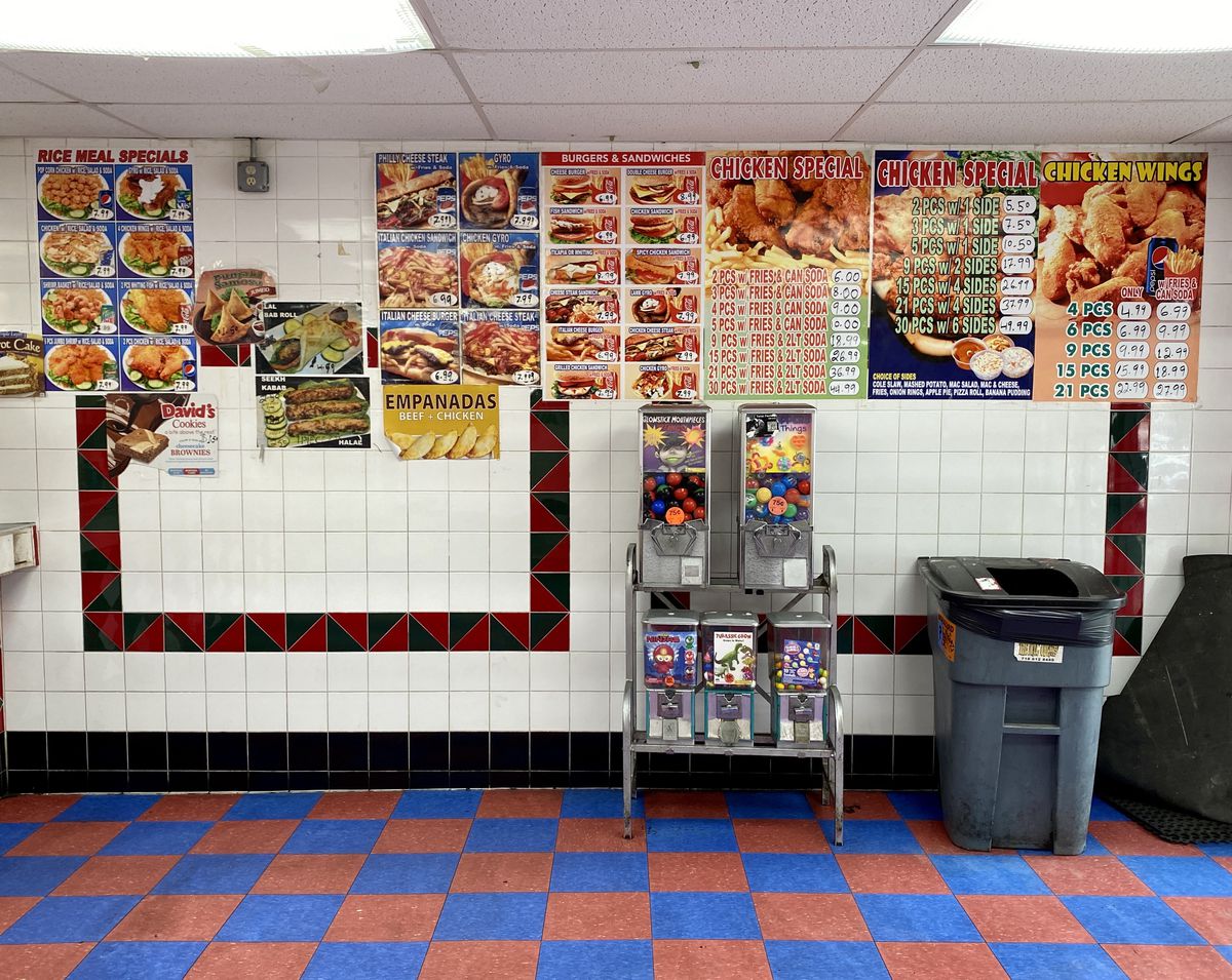 The interior of a chicken shop: blue and red checkered floor, white tile walls are plastered with large photographic menus of chicken orders, and in the foreground a series of gum ball machines and a plastic trash can.