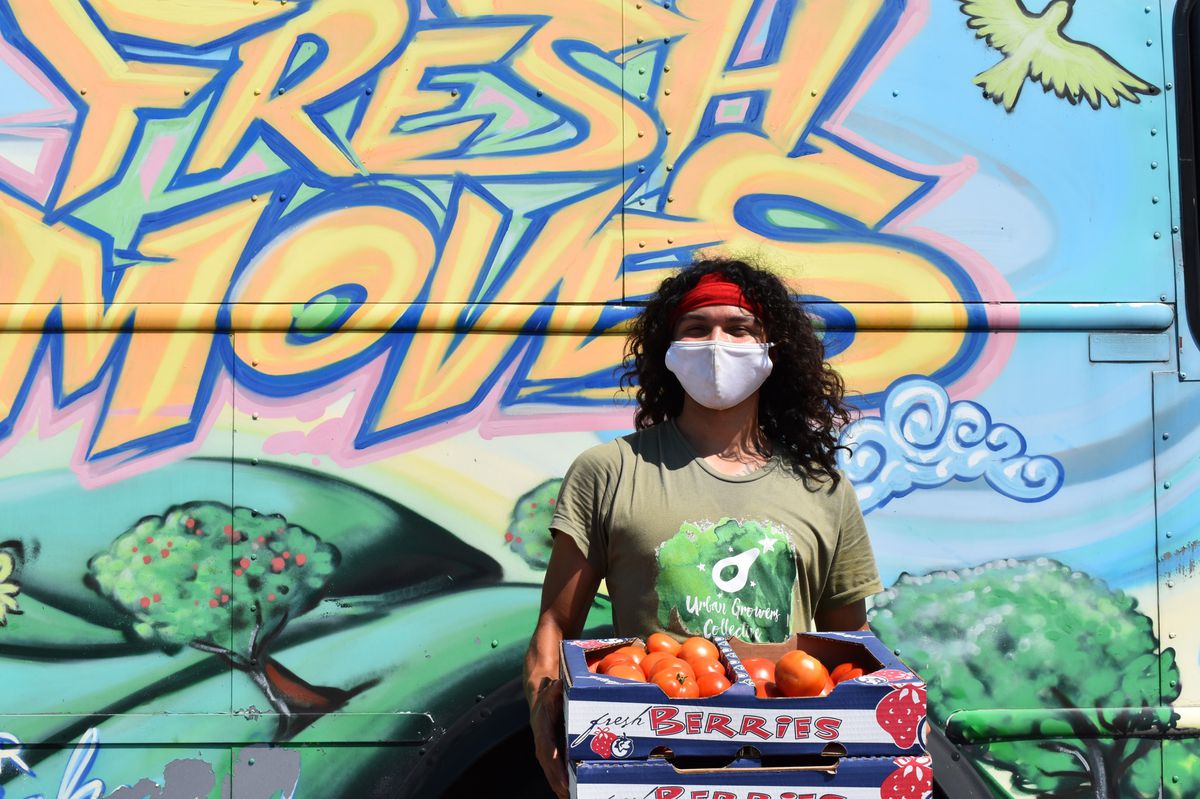 A person with wavy black hair and a white mask is holding a cardboard box of red tomatoes 