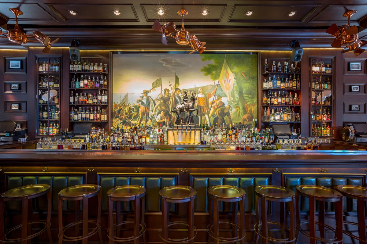 A dark-wooded bar with low stools in front and a painting of men with flags