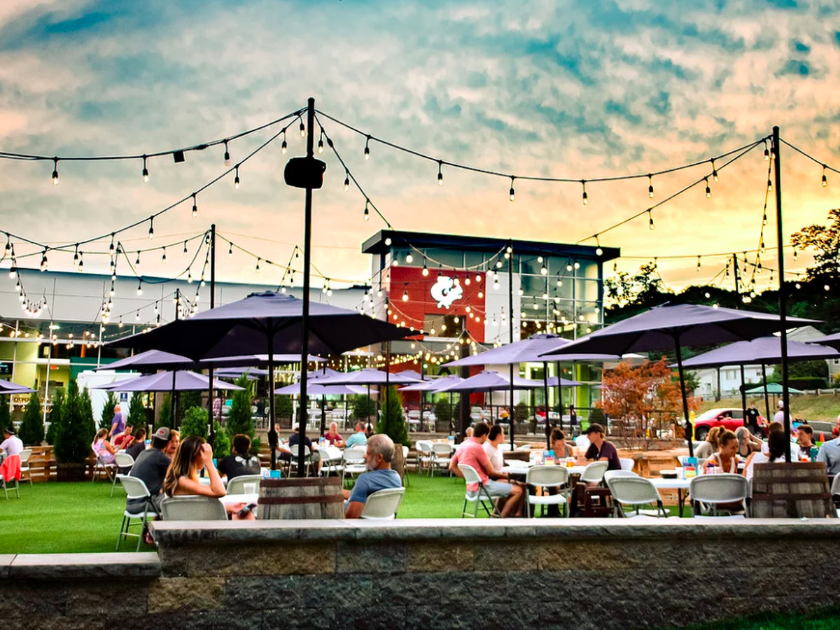 Sunset view of a large brewery patio on green turf, with blue umbrellas above each table
