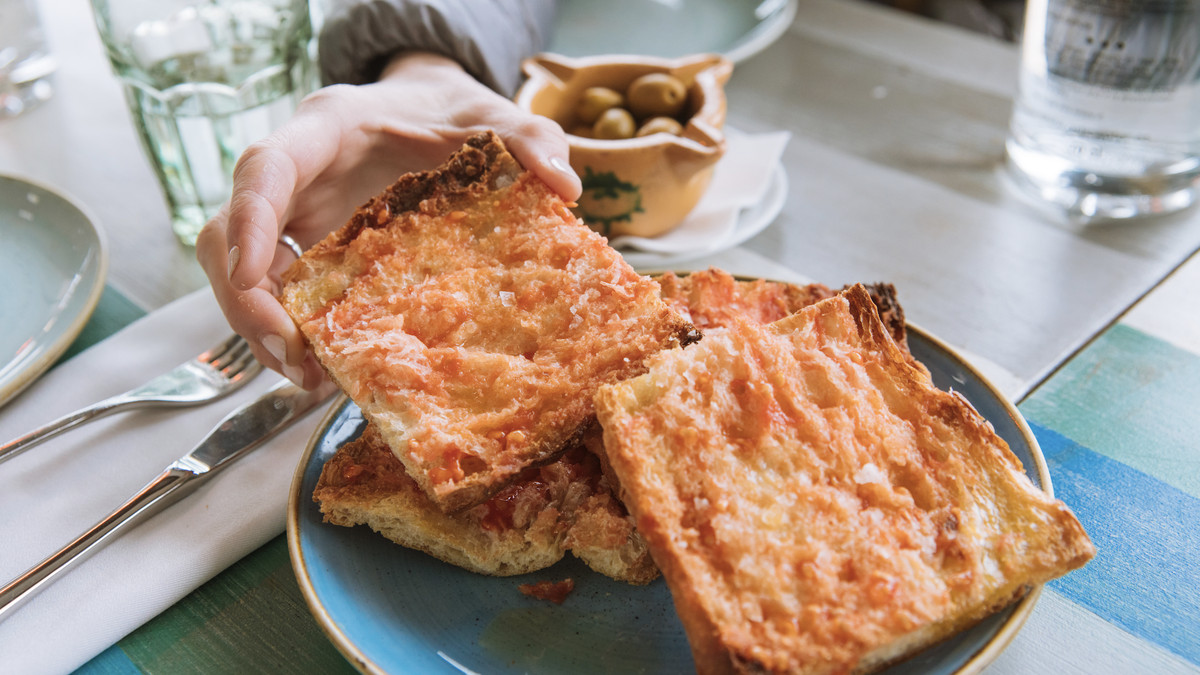 A hand reaches in to pick up a square piece of bread rubbed with tomato from a plate on a set table.
