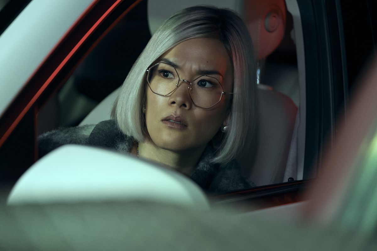 An Asian woman in octagonal-frame glasses and a silver bobbed wig sits looking out from her car.