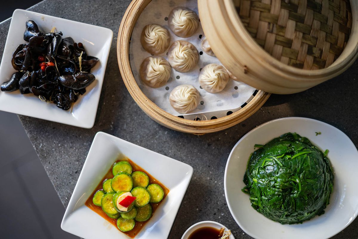 A steamer basket of delicate dumplings sits beside plates of greens, a tower of cucumbers, and a plate of wood-ear mushrooms