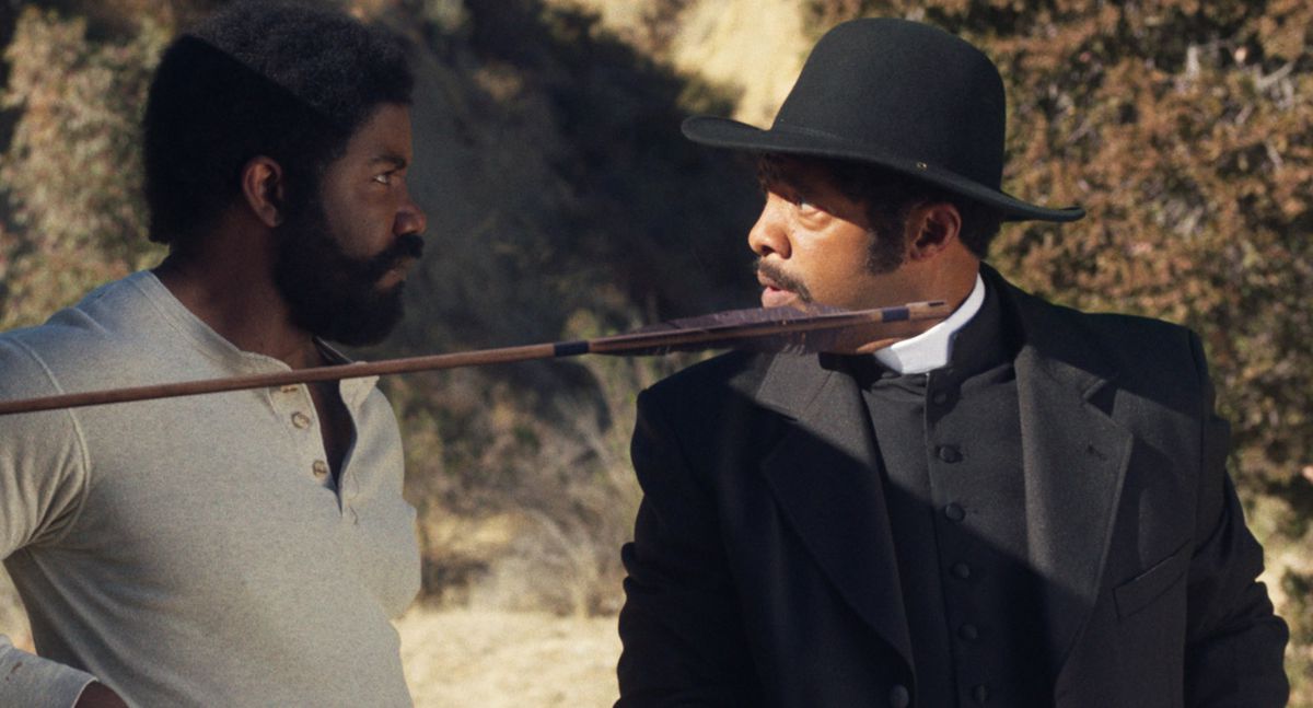 Michael Jai White and Byron Minns stare at each other in Outlaw Johnny Black. Jai White is wearing a soft top, like a part of a one-piece underwear set, while Minns is wearing preacher’s garb. An arrow sticks out in front of them.