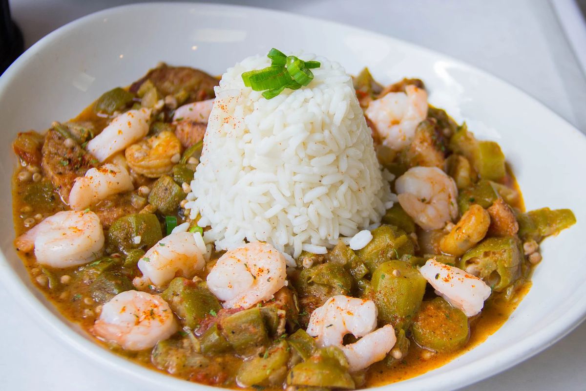 A dish with an okra, tomato, and sausage base topped with shrimp and served with white rice in the center