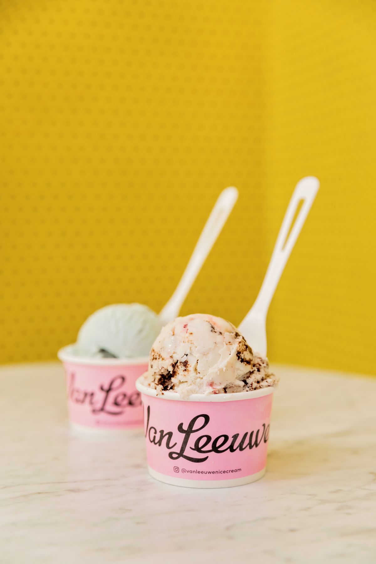 Two cups of Van Leeuwen ice cream sit in pink cups with the company logo on them. The flavors appear to be vanilla with stripes of chocolate and strawberry and mint. In the background is a yellow tile wall. The ice cream sits on a white marble counter with grey veins. 