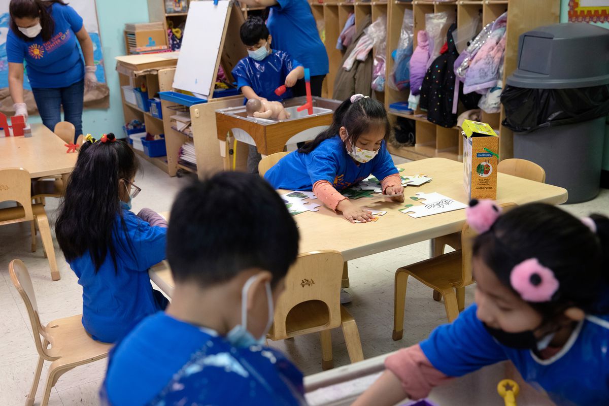 In a busy preschool classroom with young children doing various activities, a girl with black hair and a mask on fits large puzzle pieces together on a table.