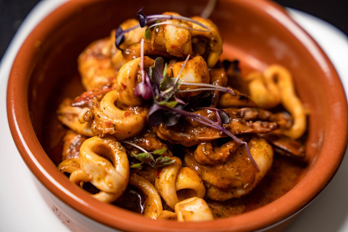 A red-brown straight-walled round tapas plate with rings of calamari covered in a red sauce and garnished with micro greens.