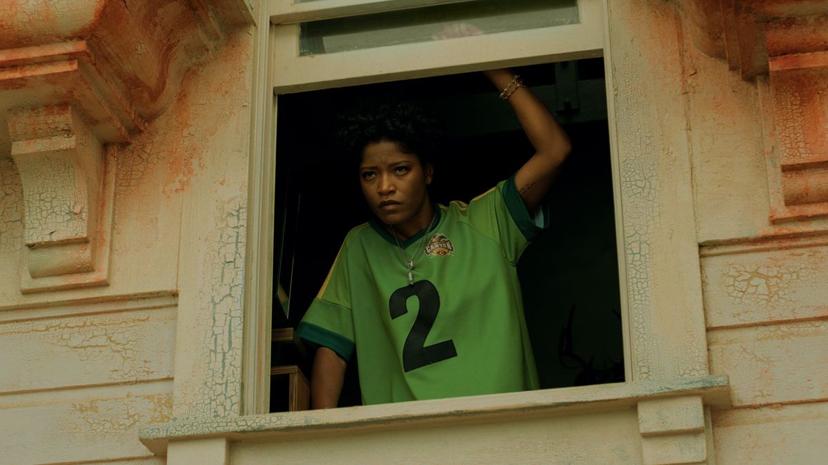 Emerald (Keke Palmer), wearing a green sports jersey, looking out the window of an old house in Nope