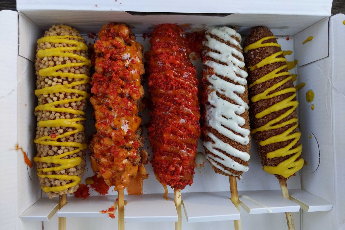 An overhead photograph of a box of five Korean corn dogs, rolled in sugar, dusted with Hot Cheetos, and coated in fried potato cubes