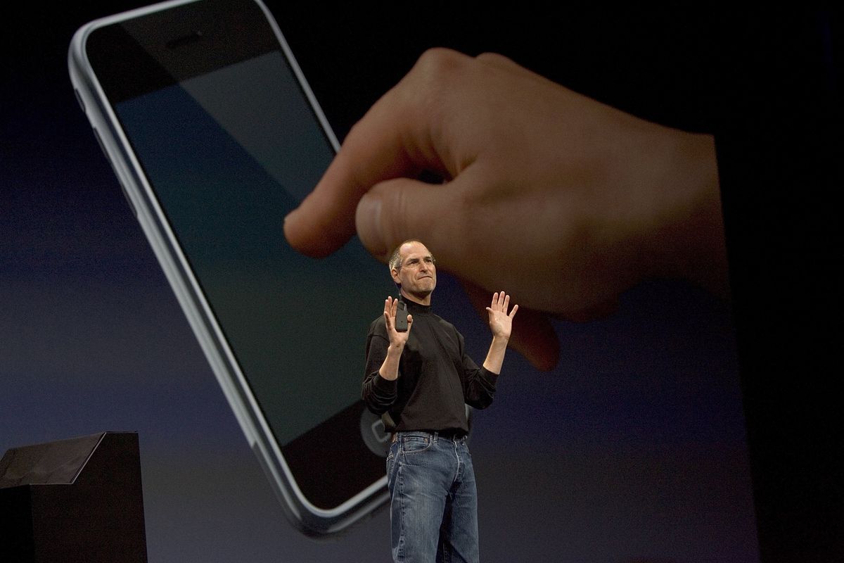 Steve Jobs onstage in front of a picture of a hand touching an iPhone screen.