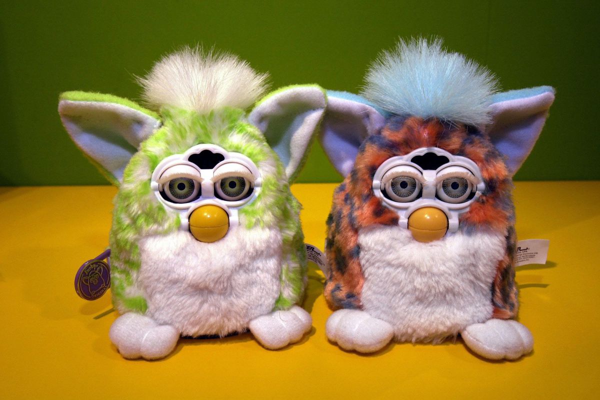 A green Furby next to a purple and orange multicolored Furby. They are on a bright yellow tabletop.
