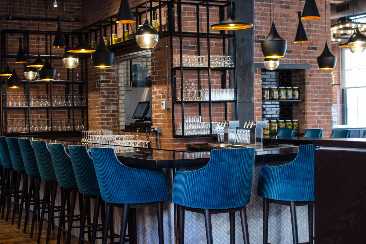 A restaurant bar in a high-ceilinged space with brick walls. The bar stools are a royal blue corduroy-like material.