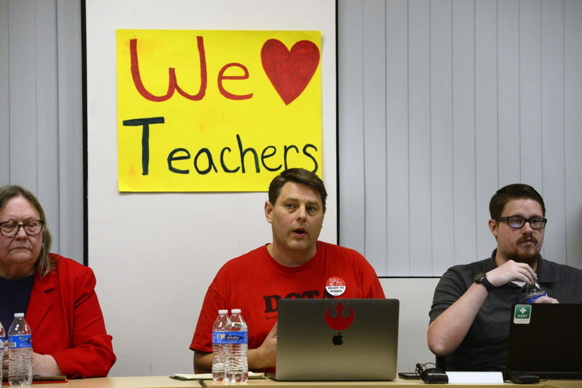 DCTA lead negotiator Rob Gould, wearing a red T-shirt, speaks as he sits between a man and a woman and in front of a We (heart) Teachers poster.