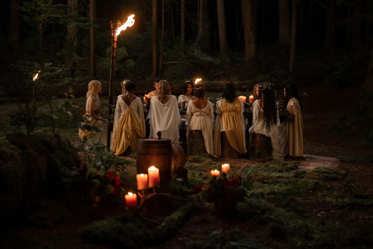 A group of teenagers in classical robes sit in the forest around a candlelit table.