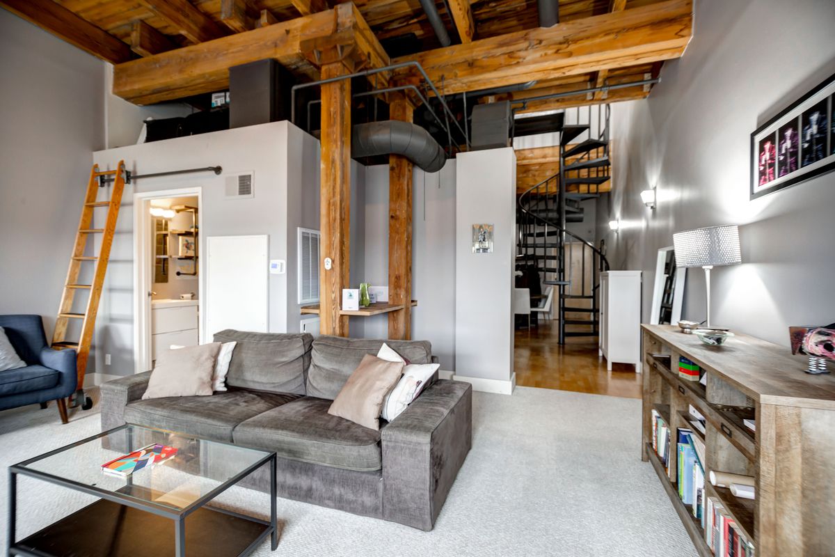 A loft with a spiral metal staircase and wood ladder leading to the second floor. On the first level there’s a gray couch and wood credenza on the carpeted floor. 