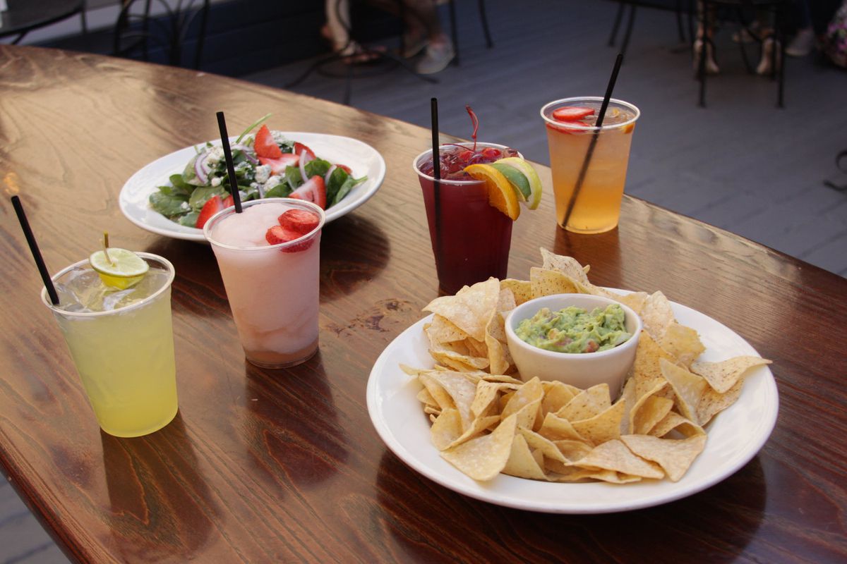 A plate of chips with guacamole sits on a table with four drinks and a salad