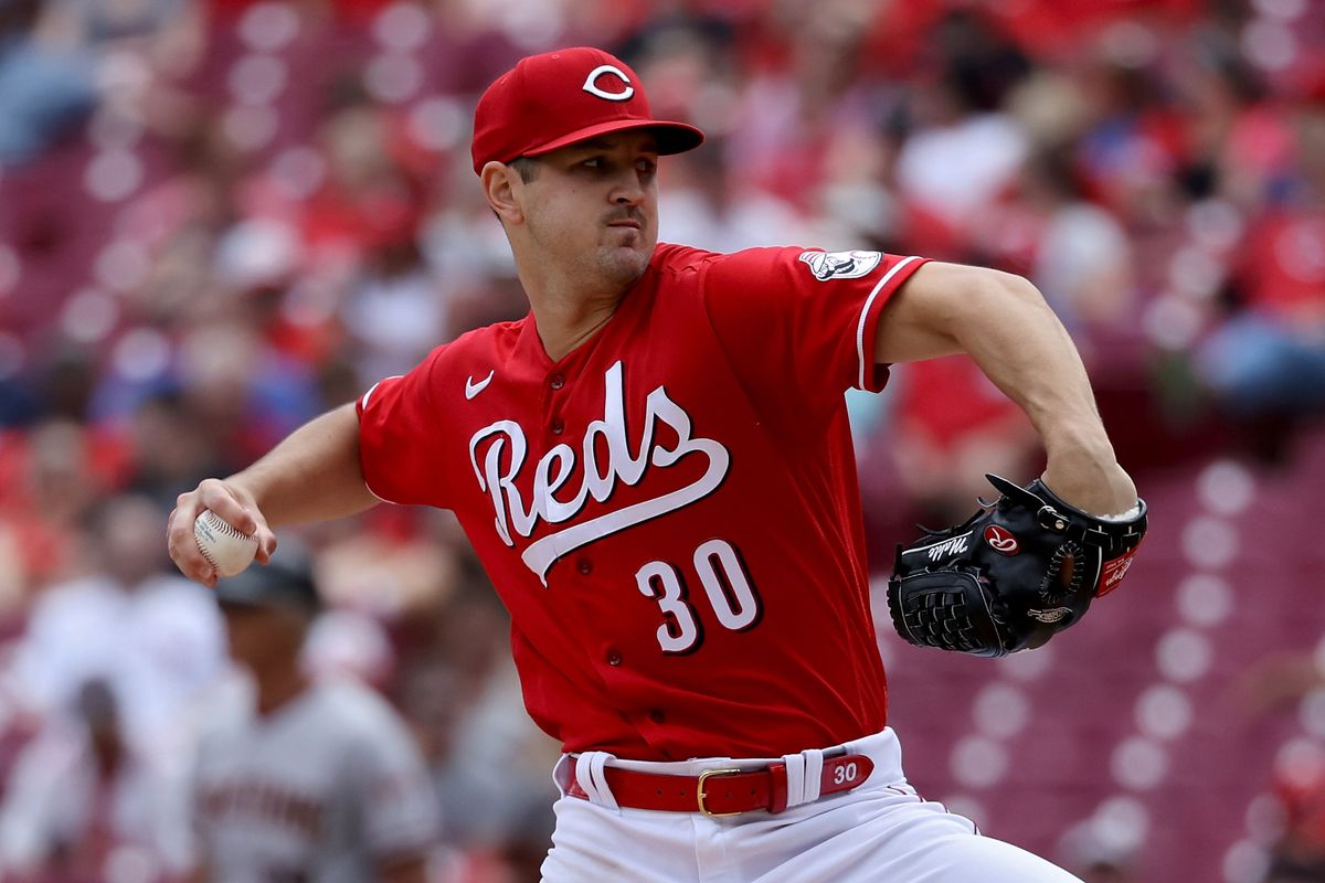 Tyler Mahle, wearing a bright red uniform with the word Reds underlined in flowing cursive, throws a pitch in front of the stands in Great American Ballpark with the crowd just out of focus.