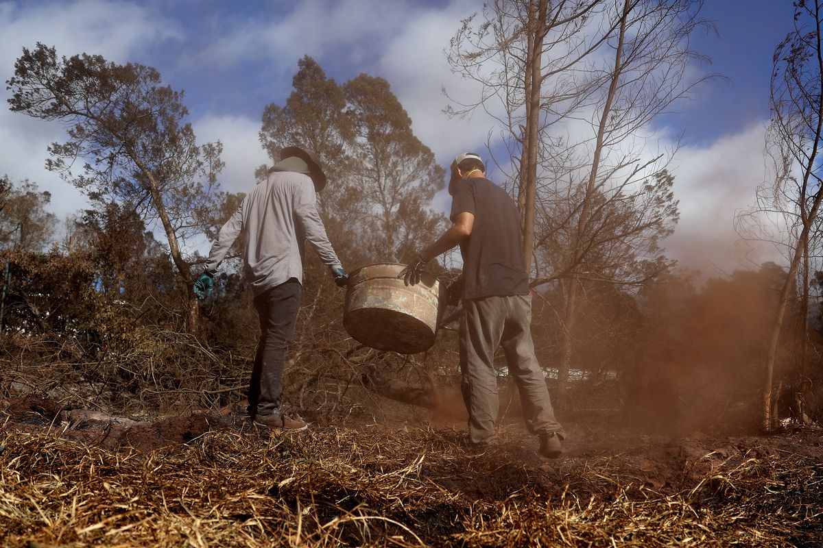 Two people stand on a patch of smoky, burned grass, holding a rusty metal bucket between them.