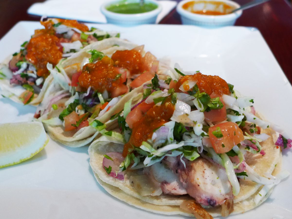 Three tacos stuffed with octopus and vegetables and sauces are linked up on a white square plate with a lime wedge on the side.