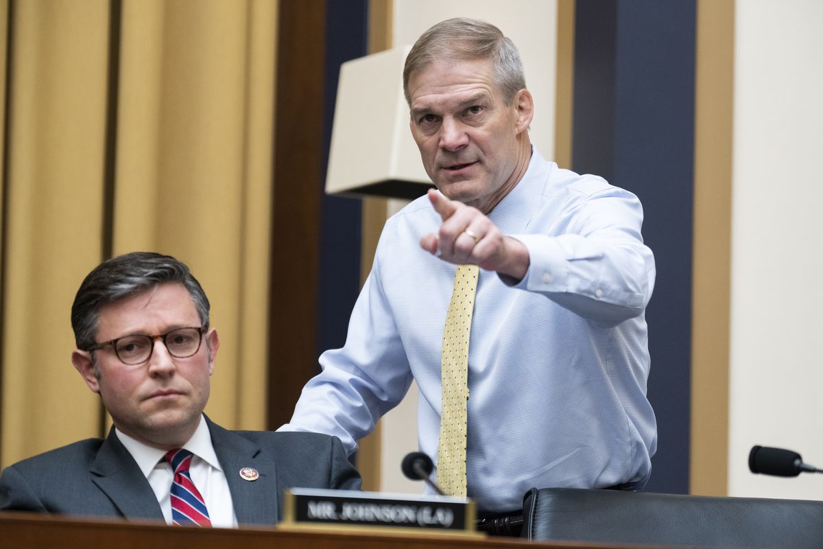 Rep. Johnson sits with a somber expression on his face. Rep. Jordan stands beside him and points a finger in the air, wearing a serious and displeased expression.