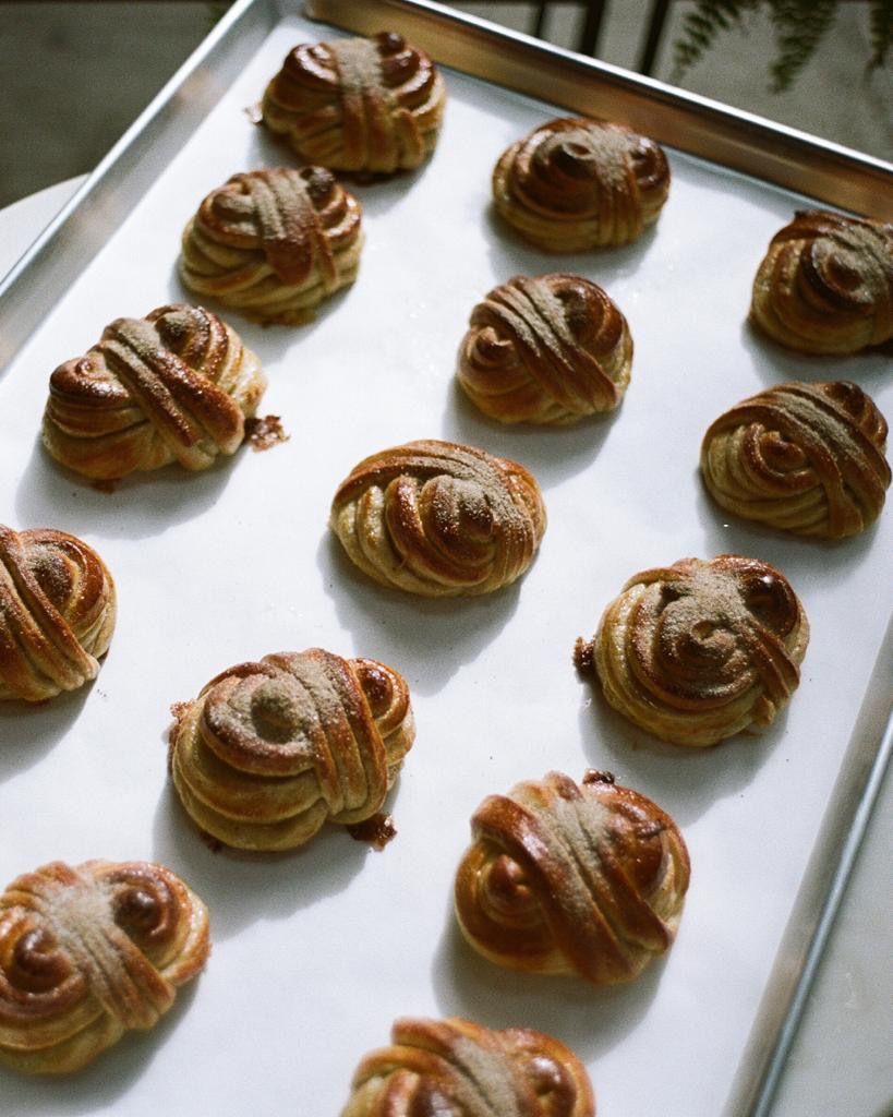 Baked cardamom buns laid out on a sheet pan and set on a white table.