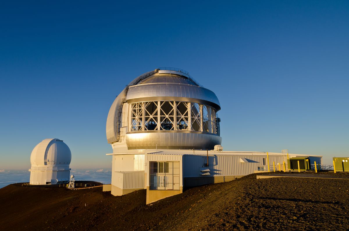 The exterior of the Mauna Kea Observatory in Hawaii.  The main building has a domed roof and a white facade.  In the distance, on the edge of a cliff, is another dome-shaped structure.