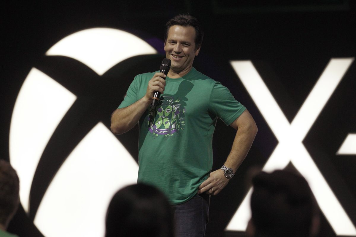 head of Xbox Phil Spencer wearing a green T-shirt and holding a microphone on stage with an Xbox logo behind him