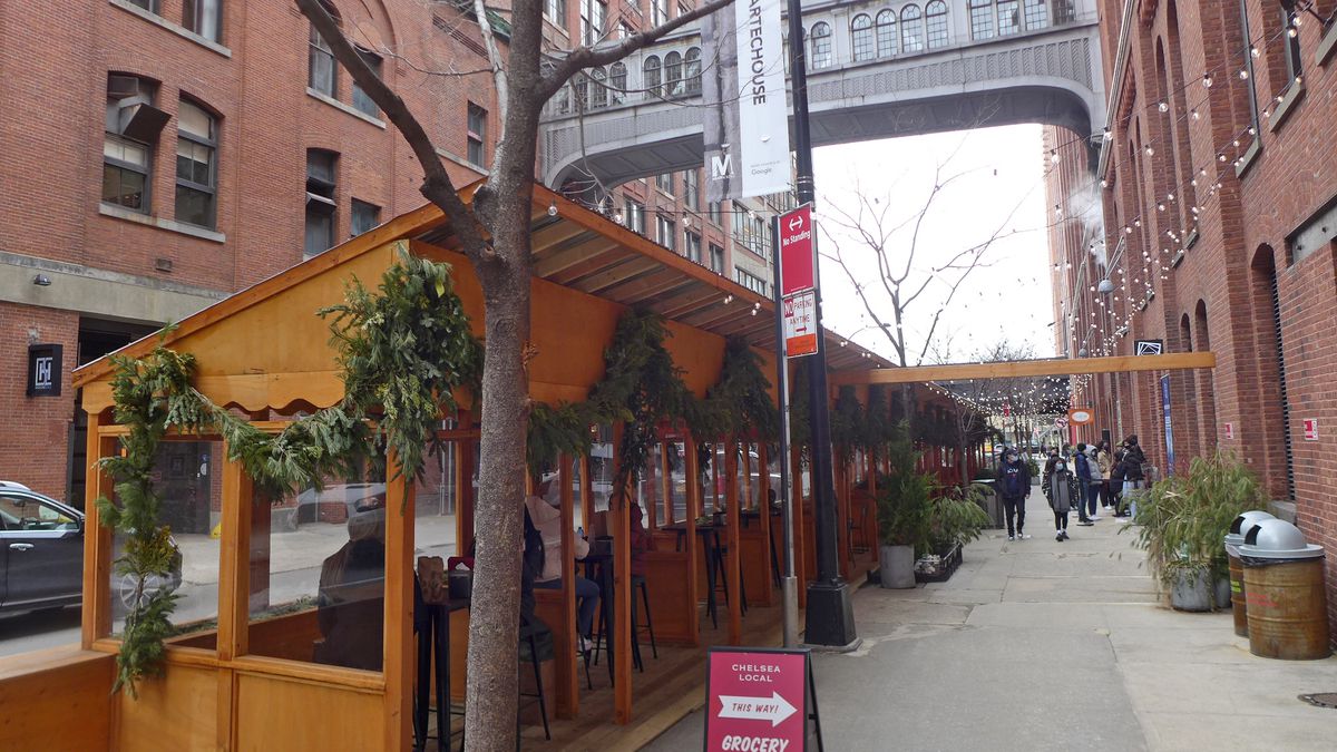 Wooden outdoor dining kiosks in foreground with bridge between two ancient brick buildings up above.
