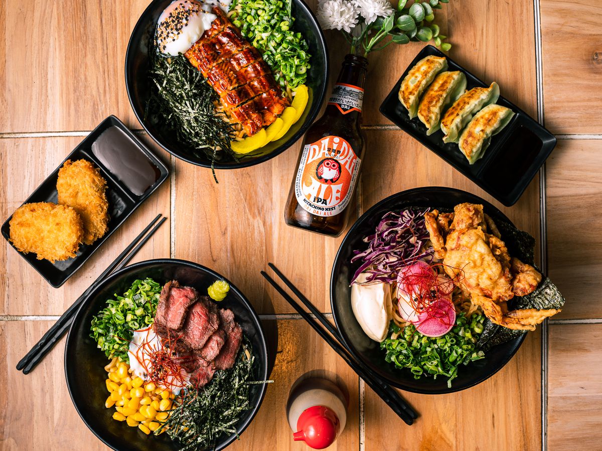 Three bowls of soba noodles with large colorful toppings, along with other dishes, surrounding a bottle of rice beer. 