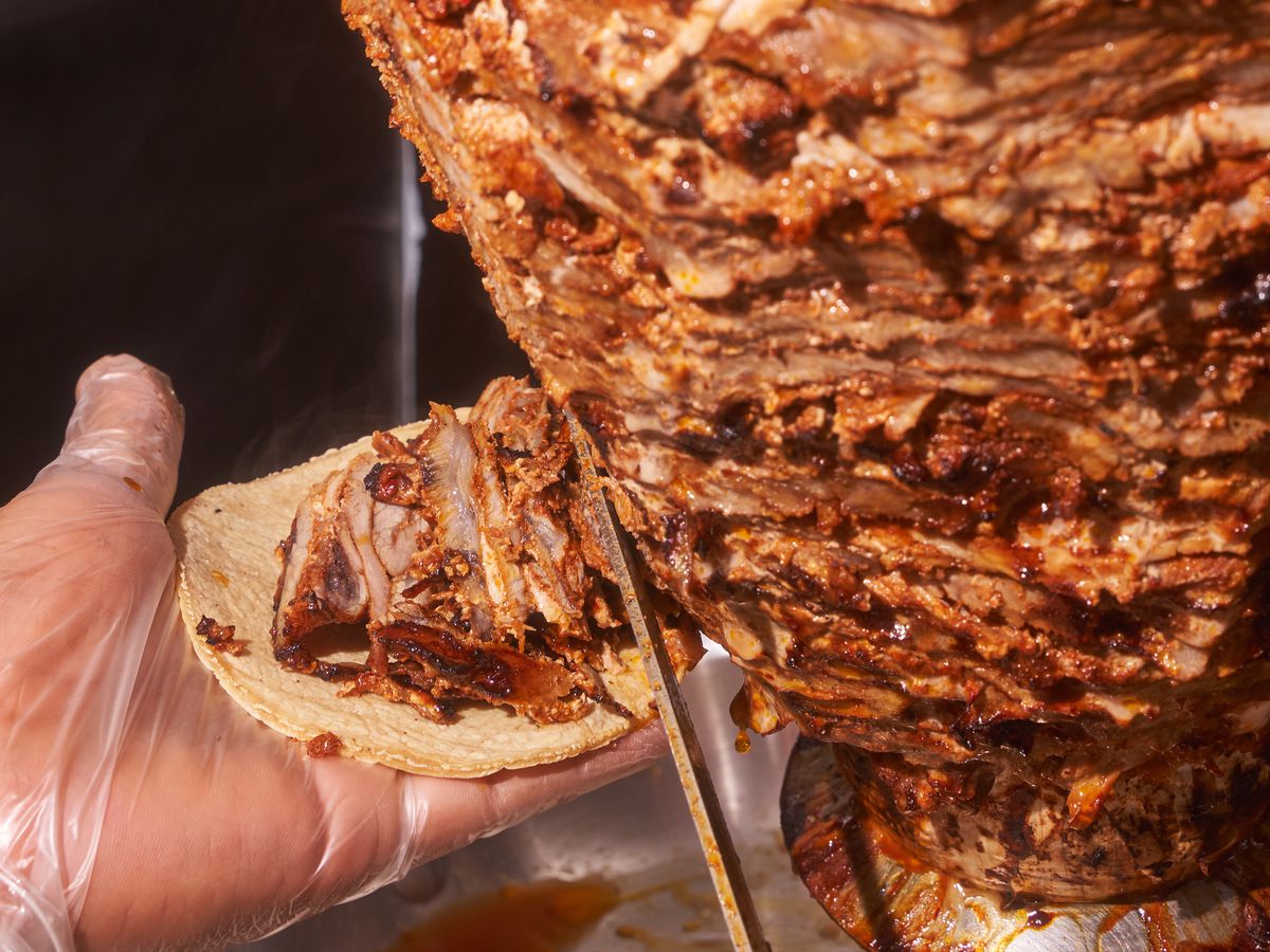 A knife cuts thin al pastor meat from the spit, which is caught by a gloved hand holding a tortilla