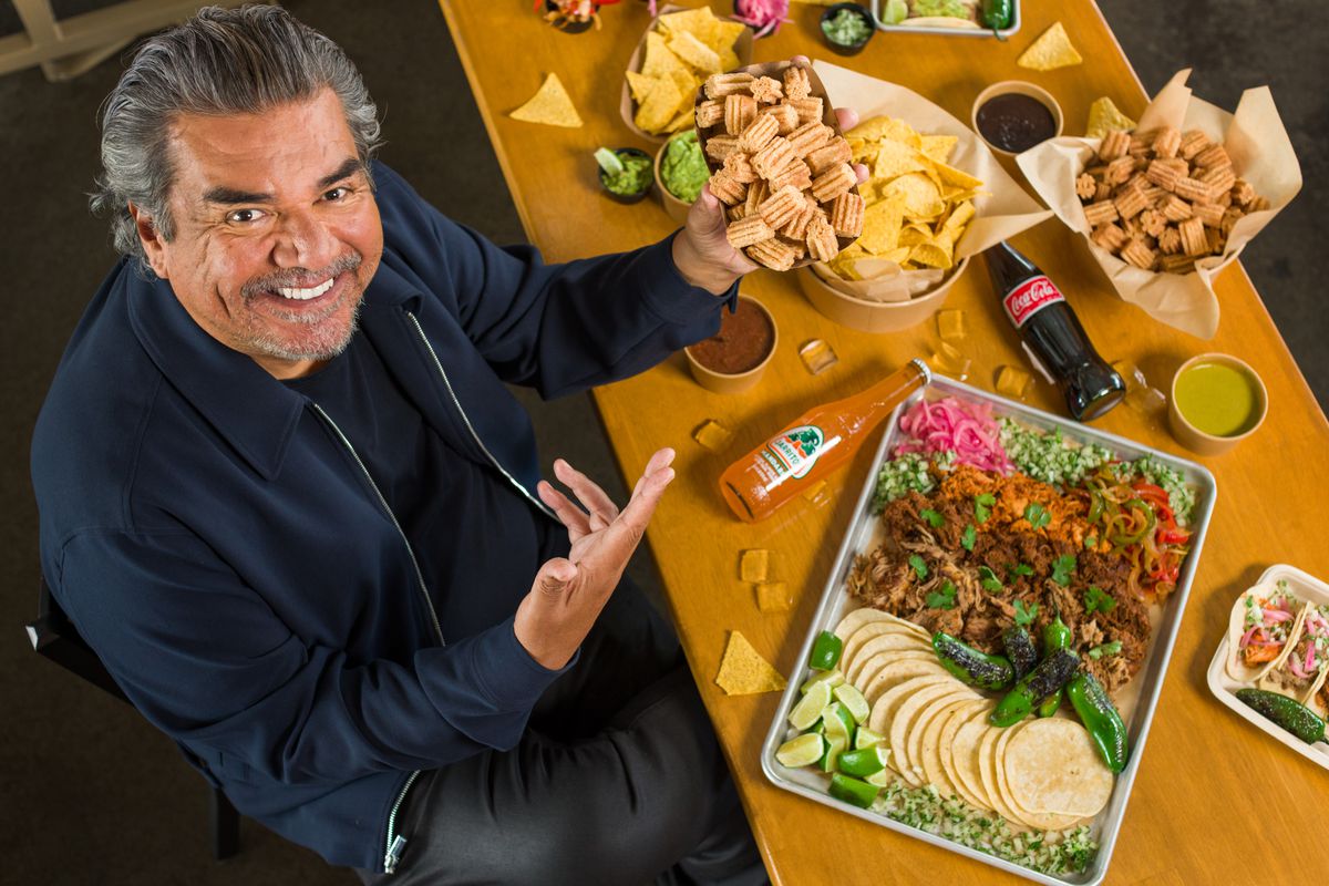 A bird’s eye view of comedian George Lopez, sitting at a table filled with tortillas, taco fillings, chips, and other sides