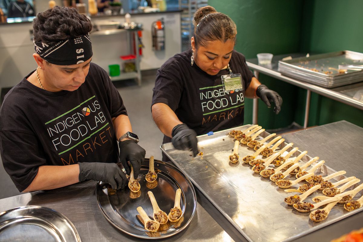 Yairany “Chiquis” Galiano and Esperanza Leal Ramos, wearing black T-shirts that say Indigenous Food Lab Market, and dishing up sampling spoons of food in an industrial kitchen. 