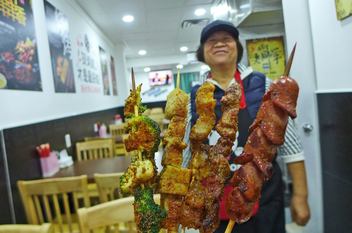 The owner, wearing a cap, stands in front of five kebabs fanned out.