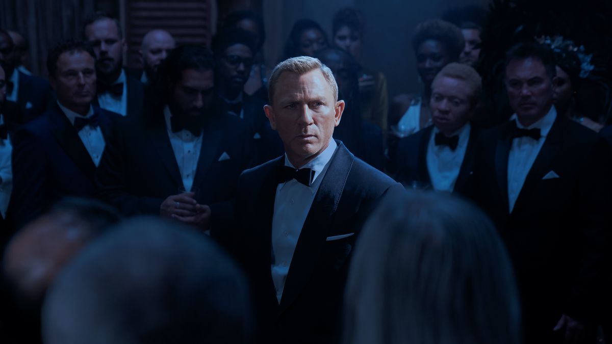 Daniel Craig as James Bond in No Time to Die, standing in a spotlight, surrounded by formal-suited members of SPECTRE