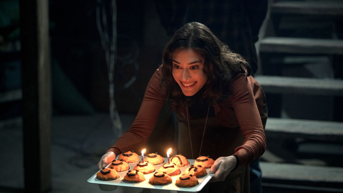 Lizzy Caplan creepily offers a tray of cupcakes, some with small birthday candles, to someone offscreen in the horror movie Cobweb