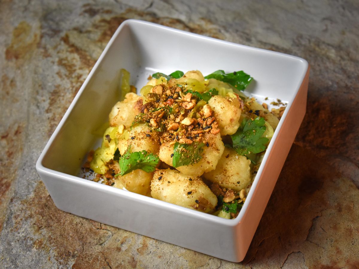 A square bowl of bright yellow cauliflower topped with chopped nuts and herbs.