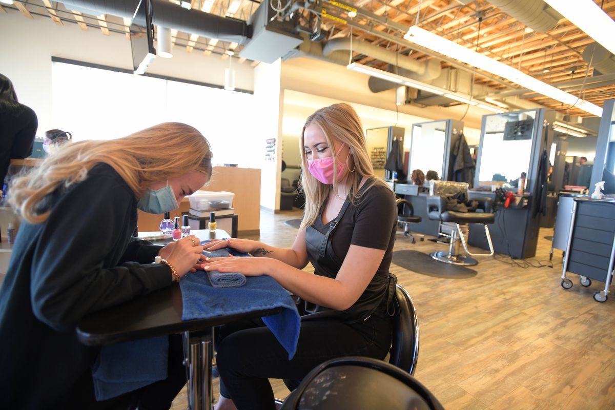 A blonde woman wearing a blue surgical mask paints the nails of another woman who’s wearing a pink mask. They are sitting at a table.