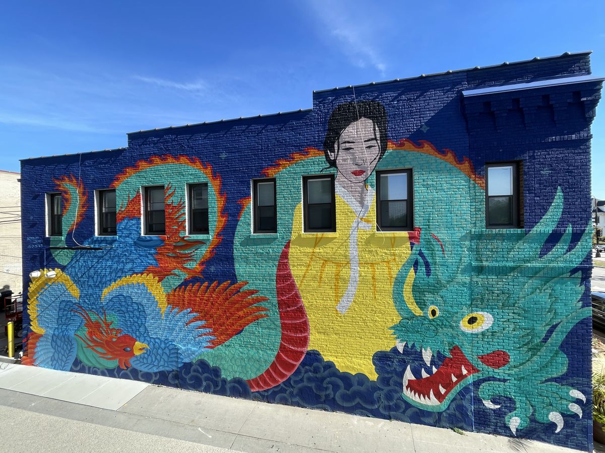 A large, vibrant mural of a Korean woman in a bright yellow hanbok intertwined with a green dragon and a blue phoenix, all on the side of a brick building.