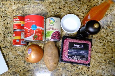 Image showing two jars of tomato sauce, a can of whole tomatoes, dark red kidney beans, salt, pepper, an onion, a potato, and two packs of “gourmet hamburger.”
