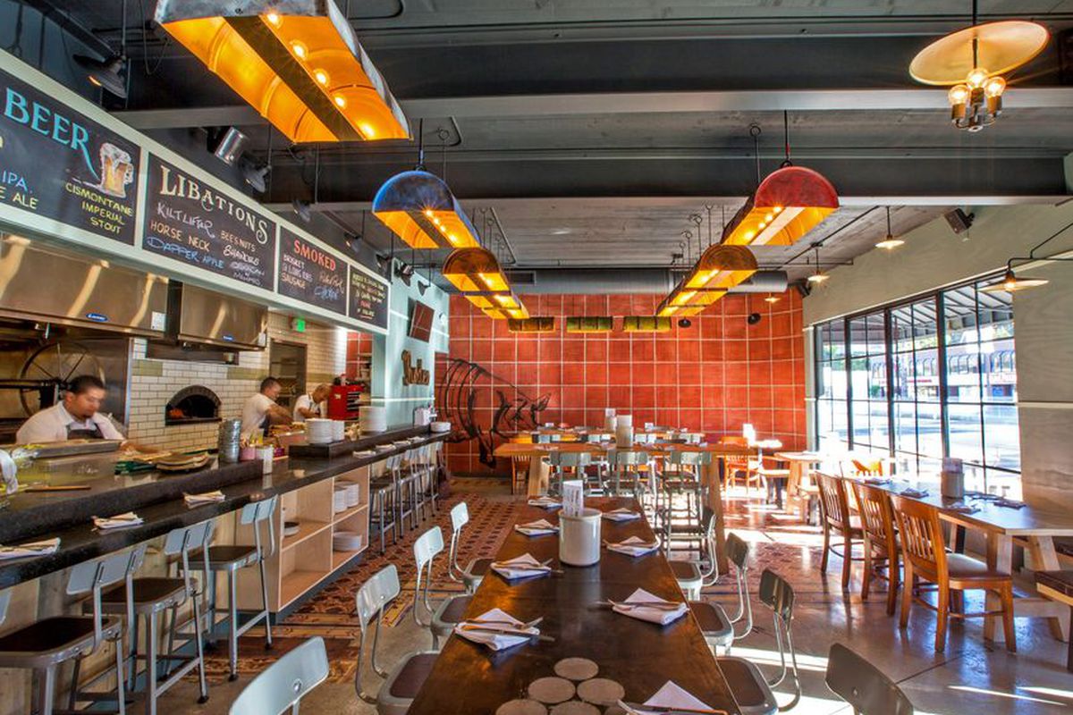 A wide look at an orange, wood-lined restaurant space with high top seats and open kitchen.