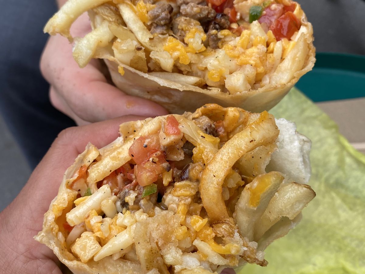 Two hands extend two halves of a California burrito, stuffed with fries, carne asada, rice, and pico de gallo